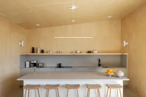A minimalist kitchen with wooden cabinets, shelves, and stools under a pendant light.