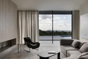 A minimalist living room with a stone floor, a grey sofa, and a black armchair, featuring a large window with view of the sky