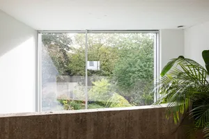Modern home interior with view of garden through large window, featuring concrete, glass, and lush green plants.