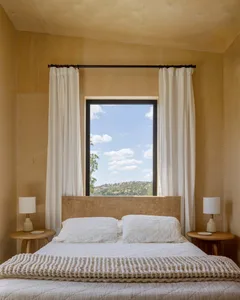 A minimalist bedroom with white curtains, a bed, and two side tables with lamps.