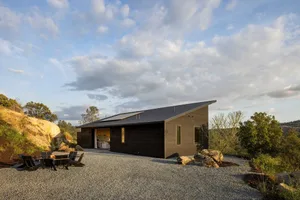 Modern residential house with wooden exterior, featuring a flat roof, large windows, and a gravel driveway with fire pit seat