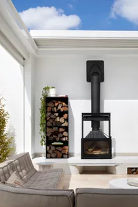 A minimalist outdoor living room with concrete and glass elements, featuring a fireplace, a sofa, and a firewood storage.