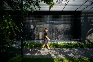 Person walking on a paved walkway bordered by greenery, with a modern black steel gate and lattice ceiling in the background.