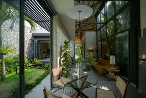 A modern interior featuring a glass table, concrete walls, and tropical plants overlooking a garden view.