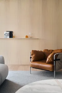 Modern minimalist living room featuring a leather armchair on a textured gray rug with a wooden wall and floating shelf.