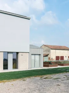A modern exterior of a building with white concrete walls, large glass windows, and a gravel driveway with green grass.