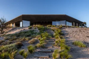 A modern house featuring concrete and glass elements on a sandy dune.