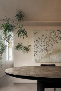 A modern living room interior with a round wooden table, hanging plants, and abstract art on the wall.