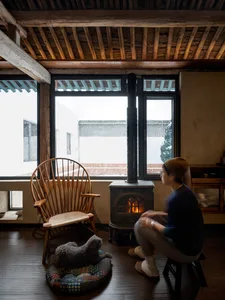 A cozy living room with a wooden ceiling, a burning fireplace, a rocking chair, and a person sitting on the floor.