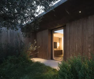 A minimalist exterior with wooden walls and a concrete roof, surrounded by greenery.