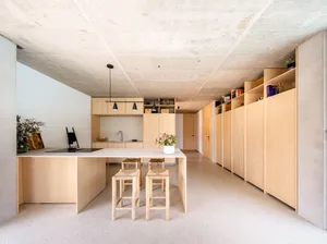 A minimalist residential kitchen with wooden cabinets, a white countertop, and concrete flooring.