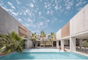 Two people jumping into a swimming pool surrounded by concrete walls with large windows and palm trees.