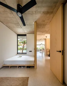 A minimalist bedroom with concrete flooring, a bed platform, and a large window overlooking a pool and palm trees.