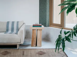A minimalist living room featuring wooden furniture, a beige sofa, a wooden side table with books and a cup, and a plant in f