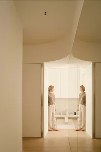 A woman standing in a minimalist residential space with beige walls and flooring, featuring geometric forms and natural light