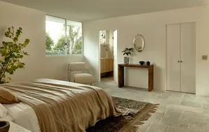 Minimalist bedroom with concrete floor, beige upholstered chair, and large window with greenery outside.