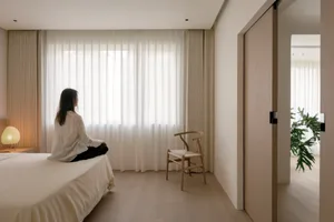 A minimalist bedroom with a white bed, wooden floor, and white curtains by a large window.