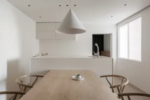 A minimalist residential kitchen with a white table, wooden chairs, and white pendant lighting.