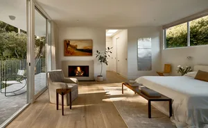 A modern bedroom featuring natural wood flooring, a comfortable white bed, and a cozy fireplace.