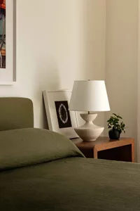 A minimalist bedroom featuring a green nightstand with a white lamp, framed artwork, and a small plant.