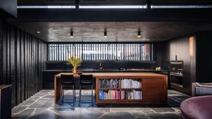 Modern minimalist kitchen with wooden island and stone flooring, featuring a bookshelf and stools.