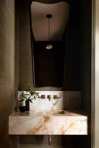 A modern bathroom interior featuring a marble countertop and backsplash, with a brass faucet and a sleek, curved mirror.