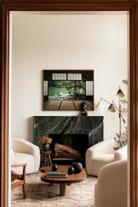 A modern living room with a marble fireplace, white upholstered chairs, a wooden coffee table, and an abstract painting.