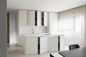 Modern, minimalist kitchen with white cabinets and countertops, featuring a sleek design and a small window.