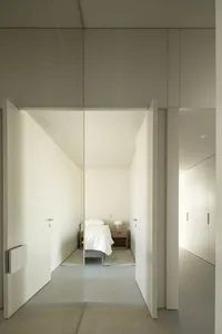 A minimalist bedroom with a single bed on a concrete floor, white walls, and a wooden bedside table.