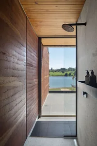An industrial-style bathroom with a wooden shower area and a view of a tranquil lake through a large glass window.