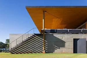 A modern exterior building with a concrete base, wood poles, and a large overhanging wooden roof providing shade.