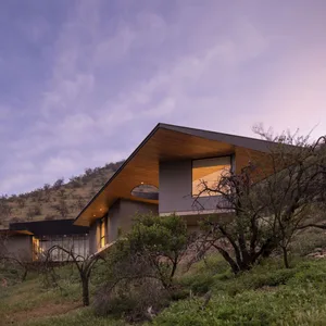 Modern house exterior with wooden roof and glass windows, set against a scenic background during sunset.