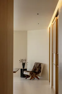 A minimalist corridor interior with neutral-toned walls and hardwood floors, featuring a single cushioned chair draped with f