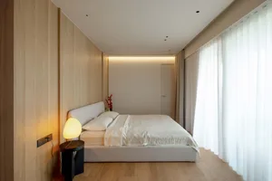 A minimalist bedroom featuring a white bed with a beige headboard, hardwood flooring, and lit floor lamp with white shade nex