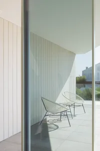 Two minimalist woven plastic chairs on a sunny patio with white walls and a glass door.