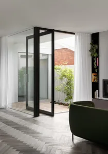 Modern living room with glass and wood elements, featuring a green armchair and white curtains.