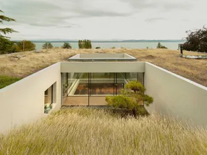 A modern house with concrete walls and large glass windows, surrounded by grass and overlooking a body of water.