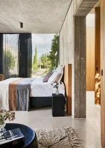 A minimalist bedroom with concrete walls, a wooden bed frame, and a large window overlooking a garden.