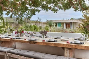 A minimalist outdoor dining area with wooden benches and cushions, featuring a long wooden table set for dinner with white pl