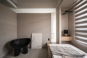 A minimalist bedroom featuring neutral-colored walls, a black chair, a bed with neutral bedding, and a wooden nightstand.