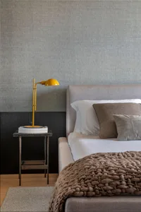 A minimalist bedroom featuring a grey upholstered bed with a taupe woven blanket, a yellow desk lamp, and a grey textured wal