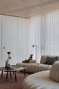 A modern sofa upholstered in soft, white textiles sits in a room with vertical blinds on the window and white walls.