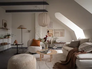 A minimalist living room with a circular wooden coffee table, a white sofa, and a fabric ottoman. The room features a round p