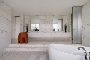 Modern bathroom interior featuring white marble walls, floor, and bathtub, with a red stool and potted plants.