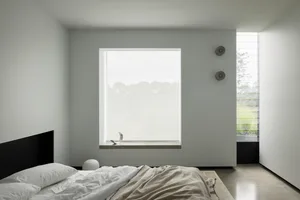 A minimalist bedroom with a concrete floor and walls, featuring a large window with a view of greenery.