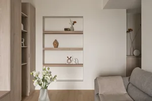 A minimalist living room featuring a gray couch, white flowers in a vase, and a floating shelf with decorative items on a neu