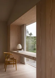 A minimalist wooden desk with a lamp and a book on a wooden floor in a well-lit room with a view of greenery outside.
