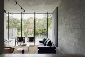 A modern living room with concrete walls, a black leather sofa, and large windows overlooking a green landscape.