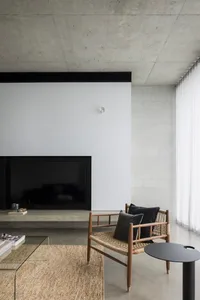 A modern minimalist living room with a canvas of concrete walls, a wood-framed chair, and a glass coffee table.