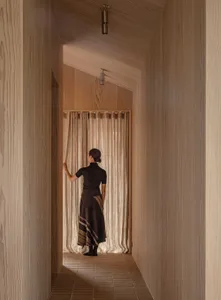 A woman standing in a corridor with wooden walls and curtains, looking through the doorway.